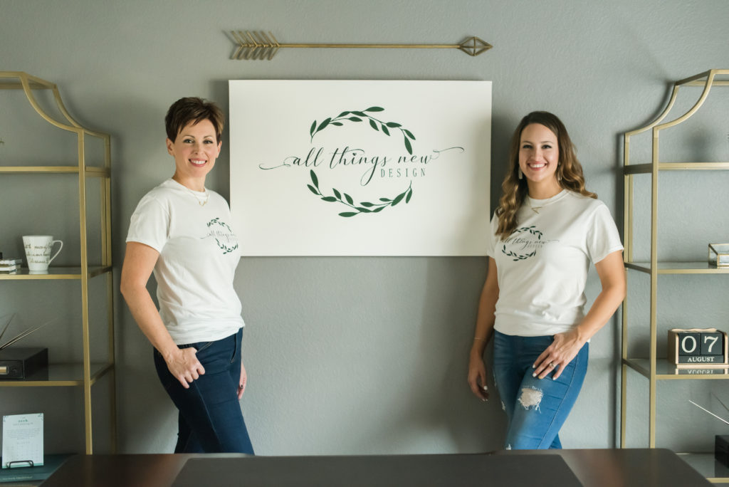Co-owners of All Things New Design in their office with a sign featuring their olive branch logo smiling at camera during photo shoot